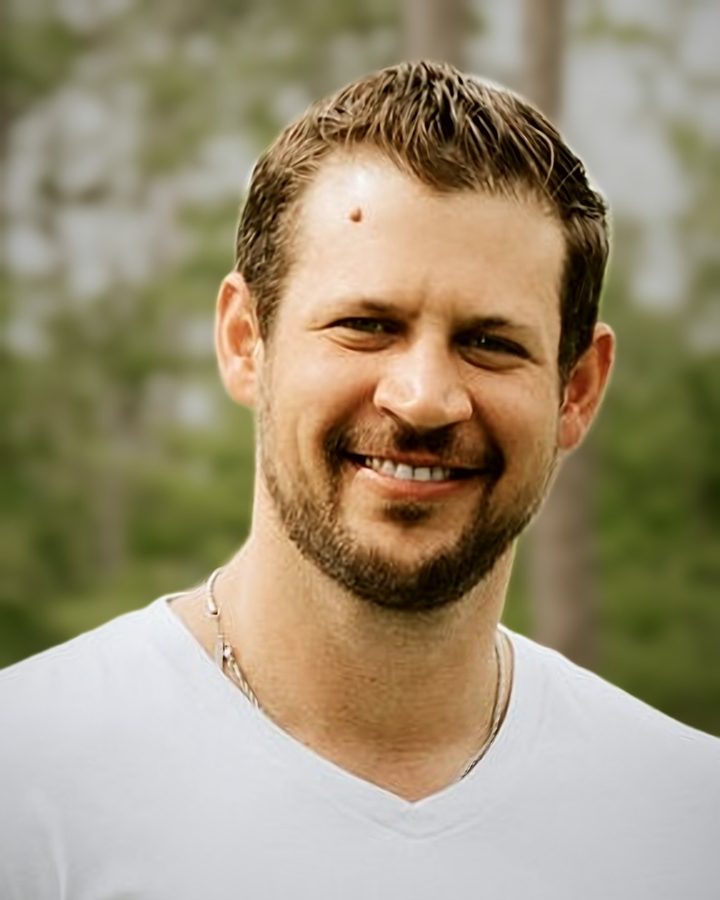Jeremy Van Winkle smiling outdoors in a white V-neck T-shirt and a thin chain necklace, with blurred green trees behind him